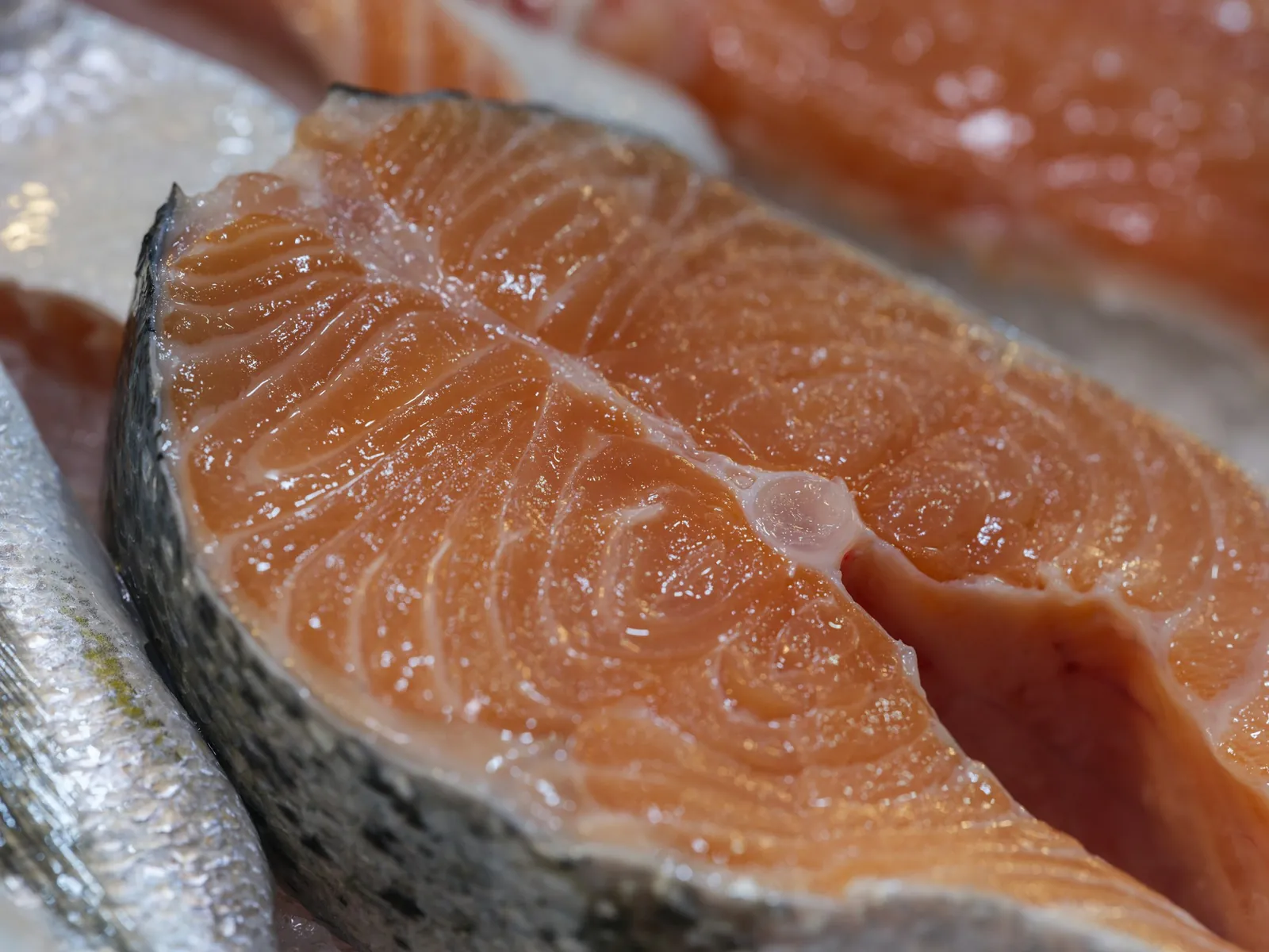Close-up of a raw salmon steak with deep orange flesh and silver skin, resting on ice beside more steaks and a whole fish