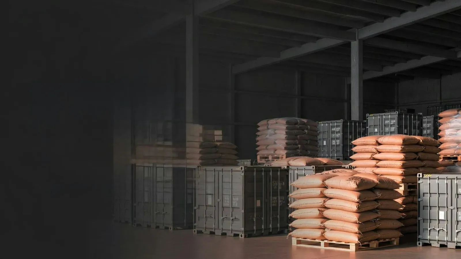 Stacks of tan feed sacks on wooden pallets beside grey metal storage containers in a dimly lit warehouse